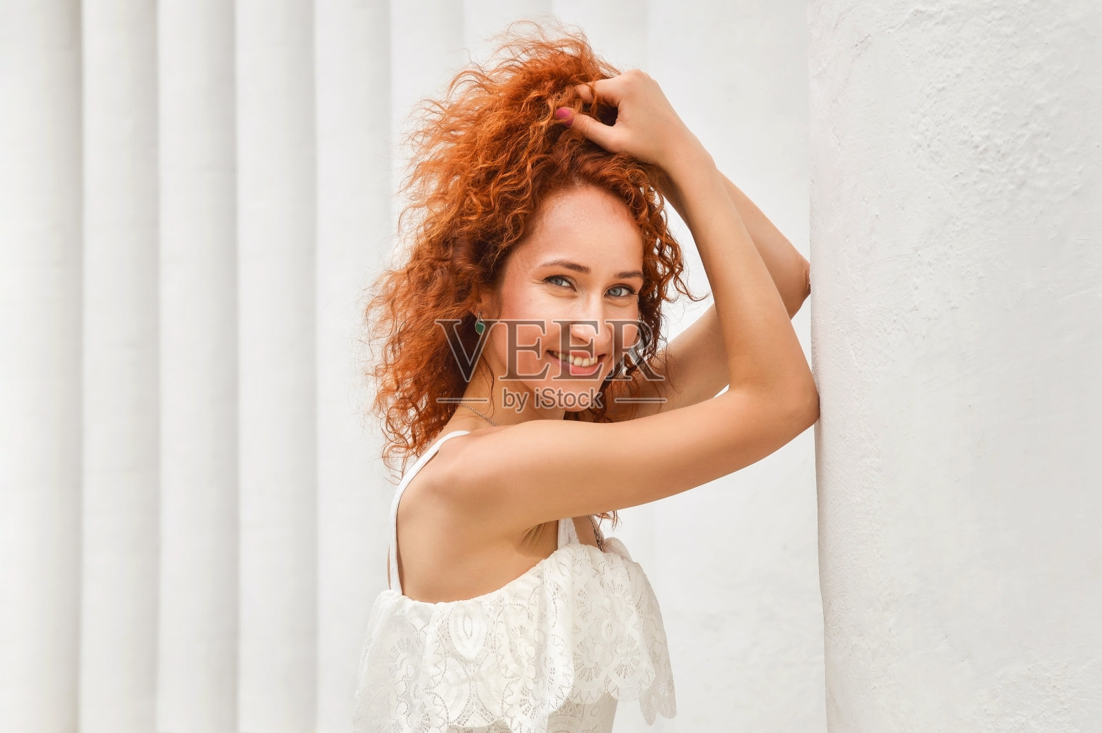 近,宏。漂亮的红发女孩笑着用手把卷发拉直。背景上的白色柱子。照片摄影图片