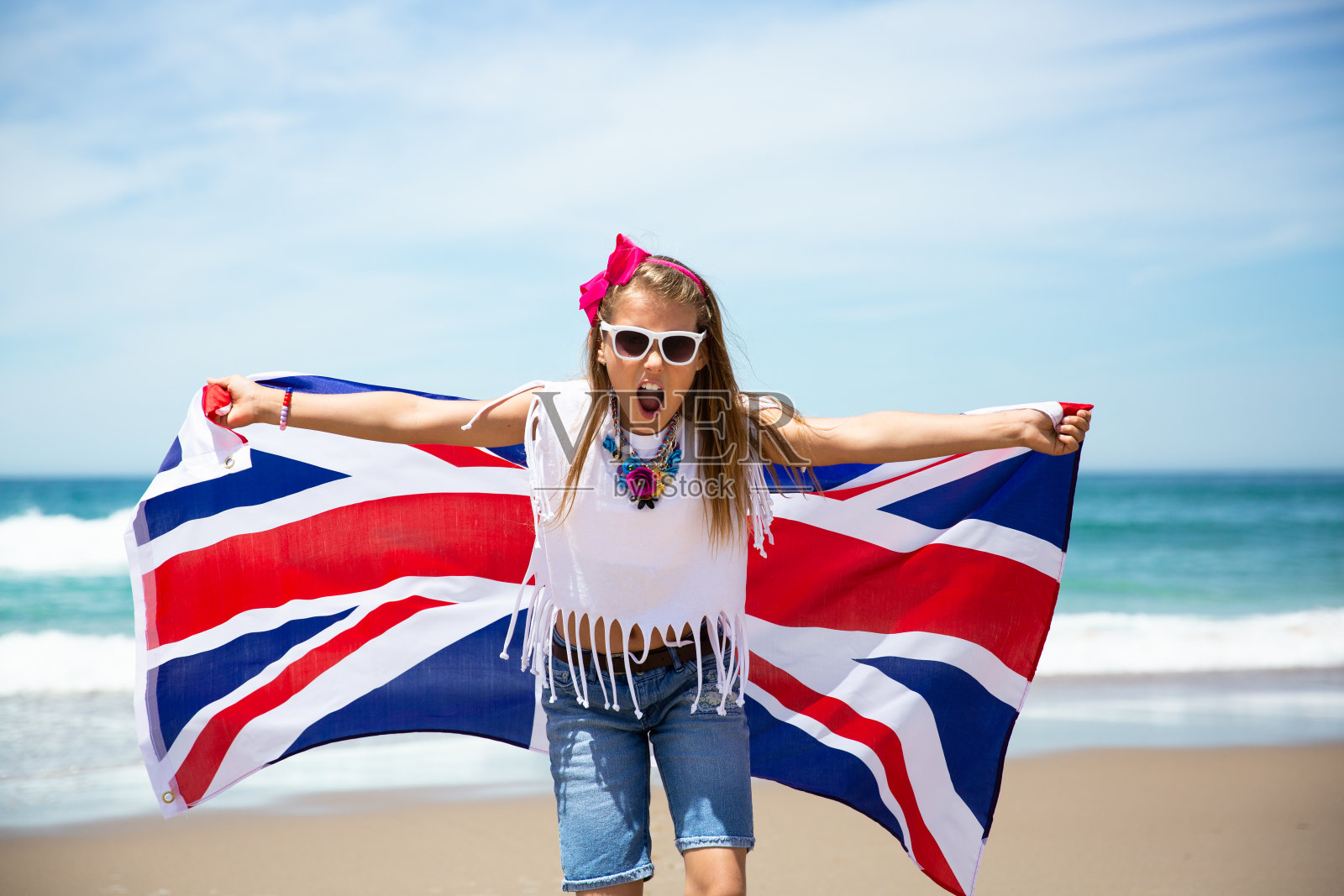 快乐的英国女孩在蓝天和大海的背景下，举着飘扬的蓝白红的大不列颠、联合王国和北爱尔兰国旗照片摄影图片