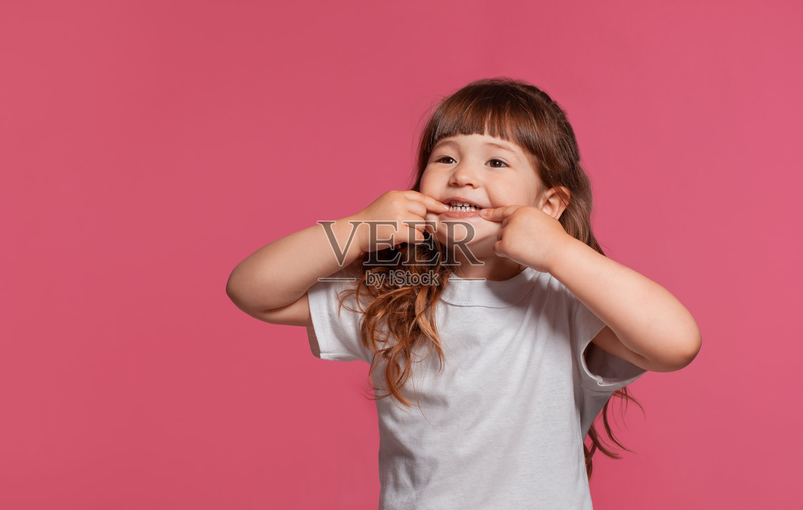 特写的一个小女孩穿着白色t恤摆姿势，以粉红色的工作室背景。真诚的情感概念照片摄影图片