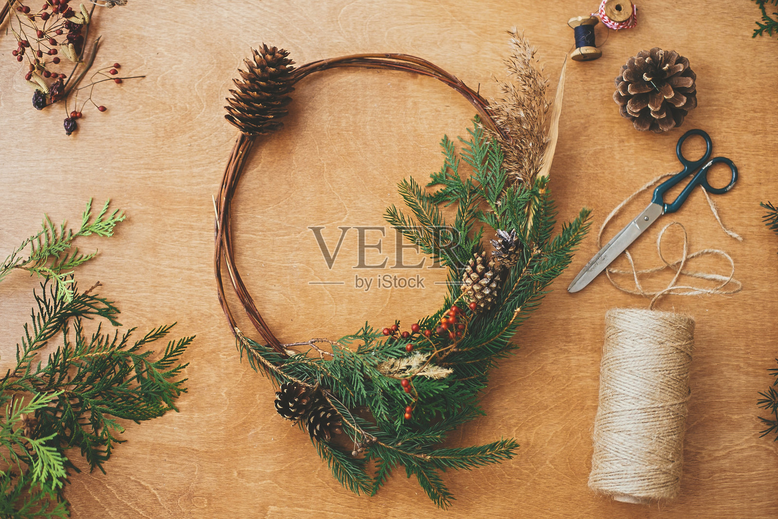 乡下人的圣诞花环，平躺着。创意圣诞花环用冷杉树枝，浆果，松果和香草与剪刀和线在乡村的木桌上。节日快乐。车间照片摄影图片