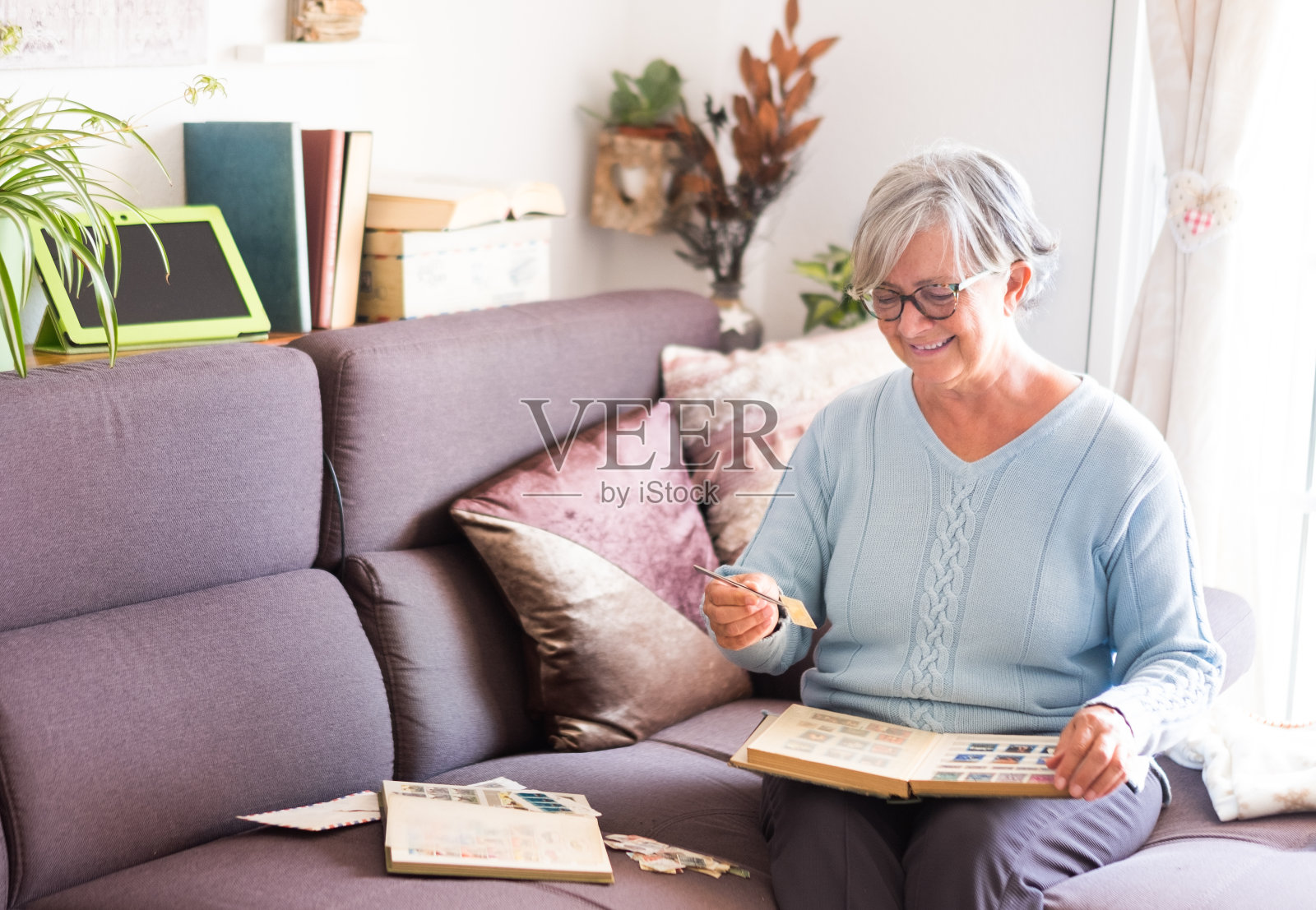 一位迷人的高级女士在看她的旧邮票集。坐在沙发上，用手机和平板电脑。白色的墙壁和书的背景照片摄影图片