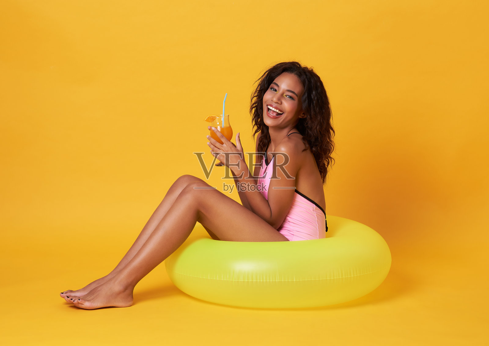 快乐的年轻非洲女人穿着泳衣举着橙汁和橡胶环在海滩上摆姿势。照片摄影图片