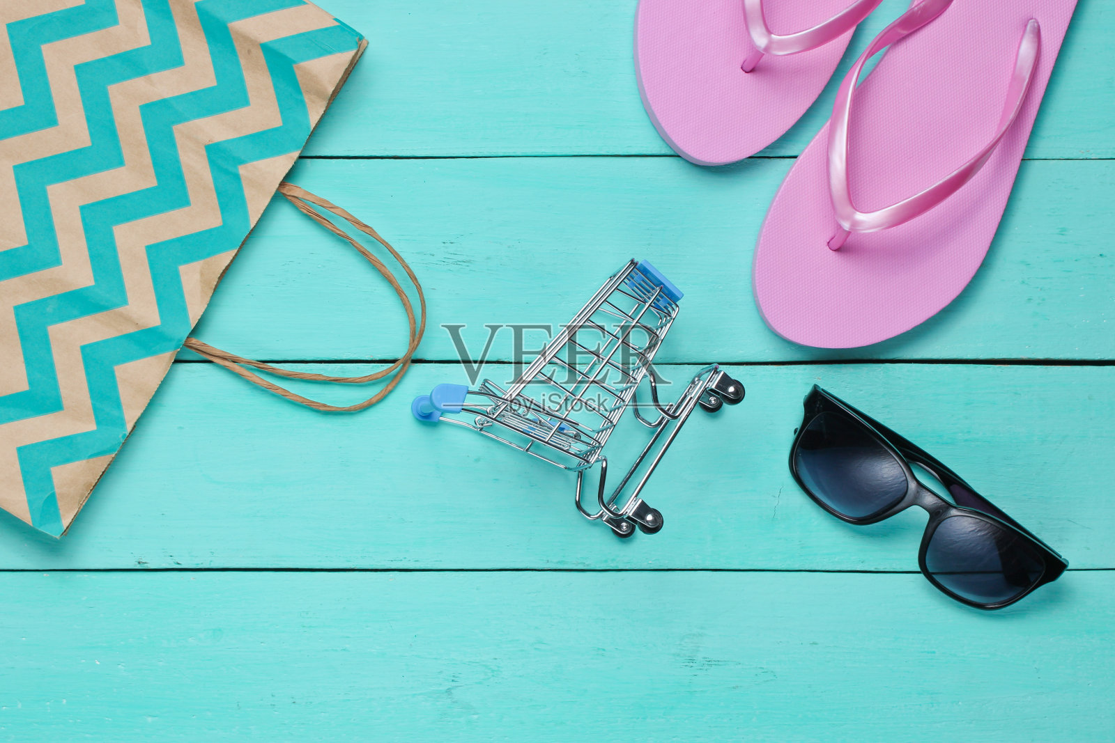 夏季出售。购物的概念。夏日沙滩饰品(人字拖，太阳镜)，蓝色木制背景的迷你购物车。前视图。平躺照片摄影图片