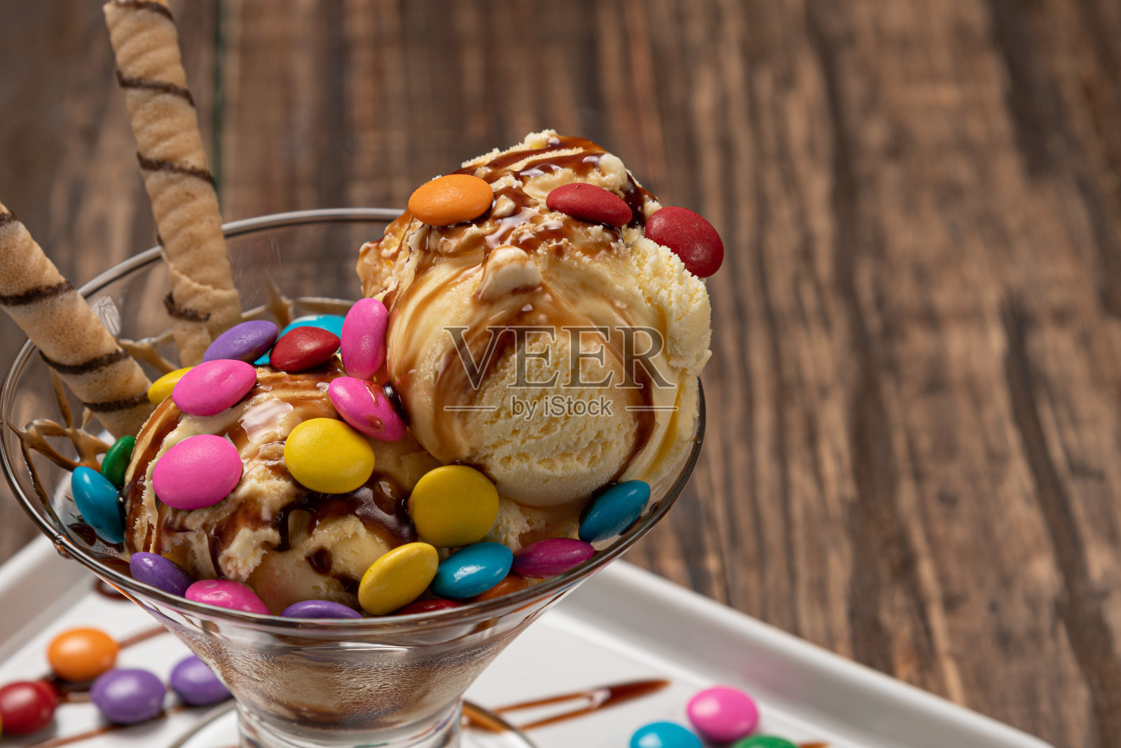 香草冰淇淋圣代玻璃碗与卷威化棒和巧克力酱在木制背景照片摄影图片