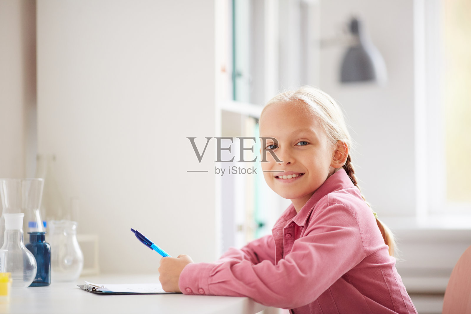 微笑美丽的女学生的肖像与金发辫子坐在桌子在实验室和做实验笔记照片摄影图片