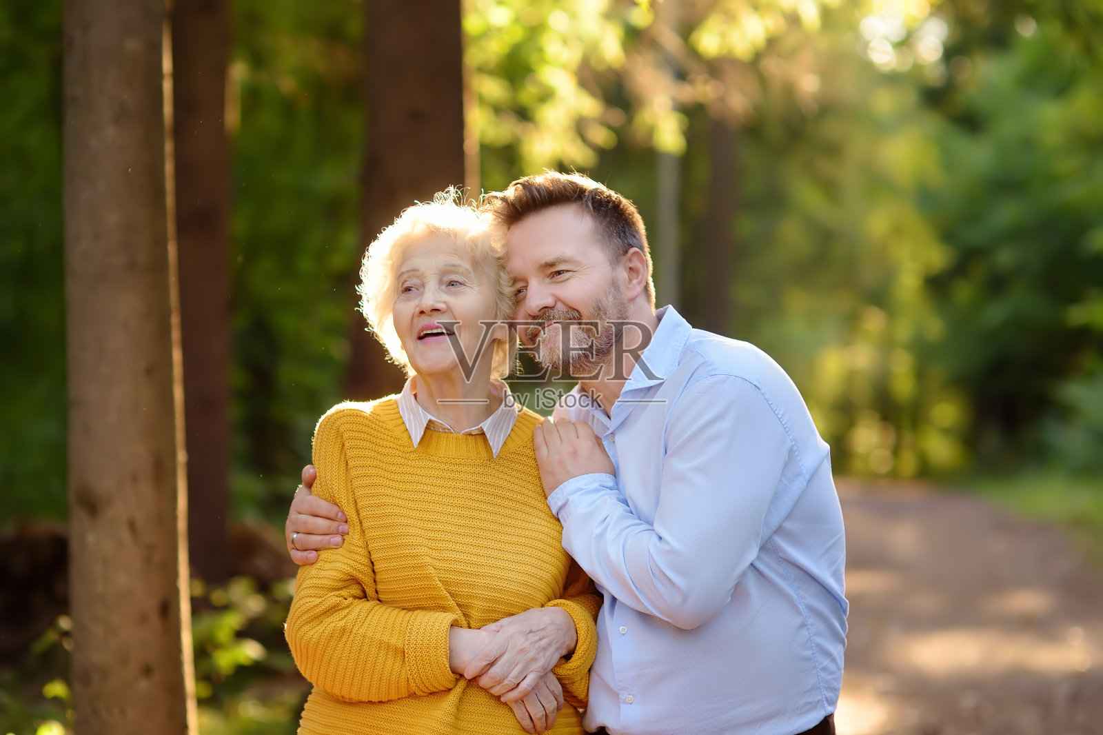 在夏季公园散步时，慈爱的成年儿子温柔地拥抱他快乐的老母亲。照片摄影图片
