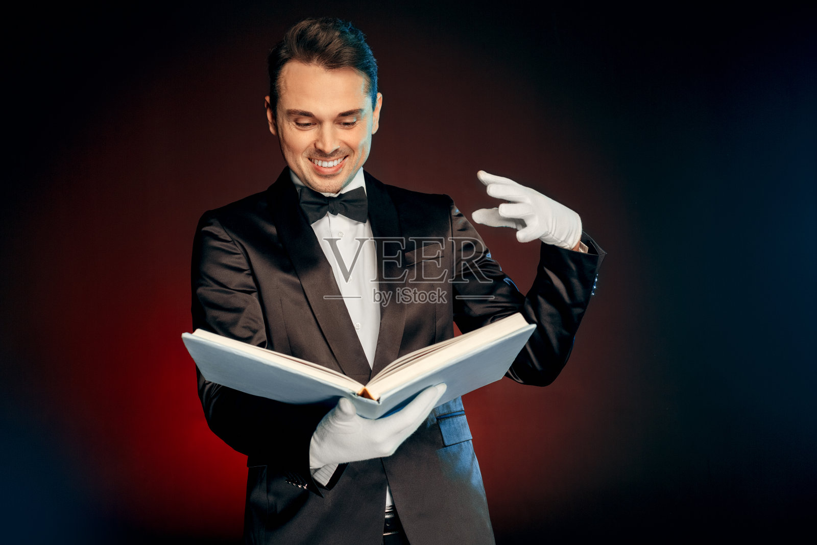 专业的职业。魔术师穿着西装和手套孤立地站在墙上，用魔法书笑得很兴奋照片摄影图片