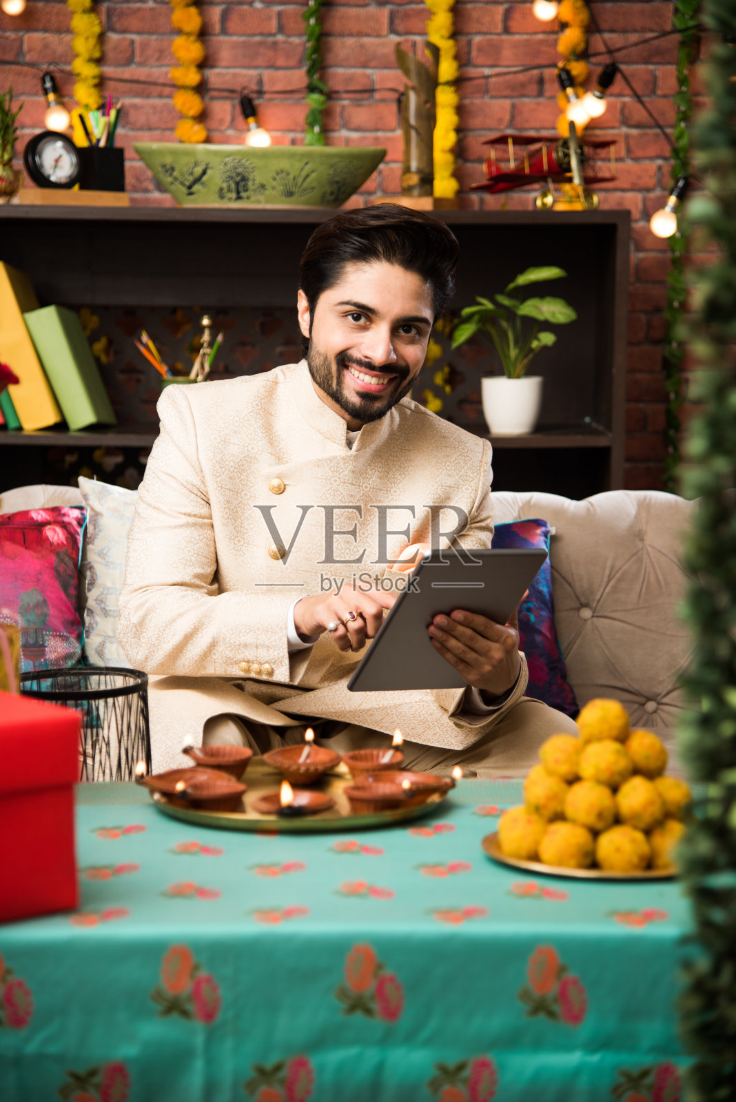 印度男子在节日当天穿着传统服装使用平板电脑。坐在沙发上，带着礼物和糖果，带着灯饰散焦照片摄影图片