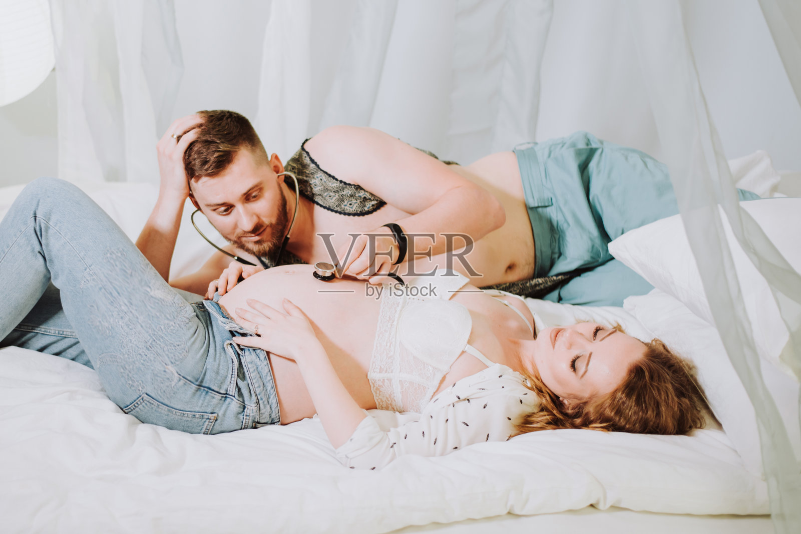 一个年轻人带着热情、喜悦和惊奇的心情，用听诊器听着他美丽的怀孕妻子裸露的肚子。照片摄影图片