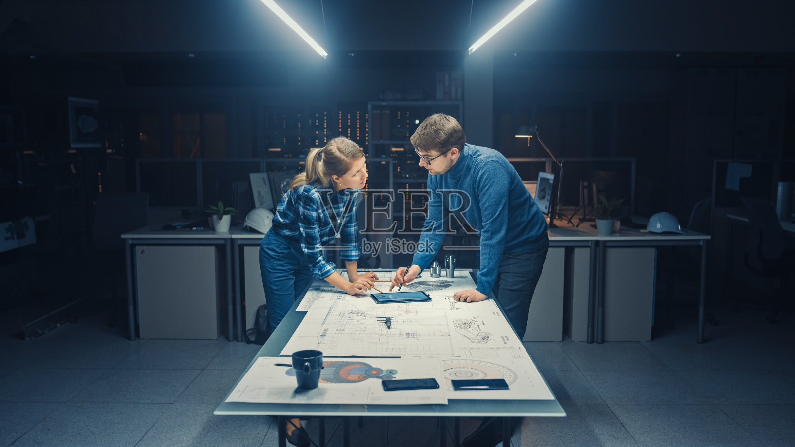 在黑暗的工业设计工程设施中，男性和女性工程师使用会议桌谈论和工作的蓝图。在桌面上的图纸，草案和电动发动机部件，零件照片摄影图片