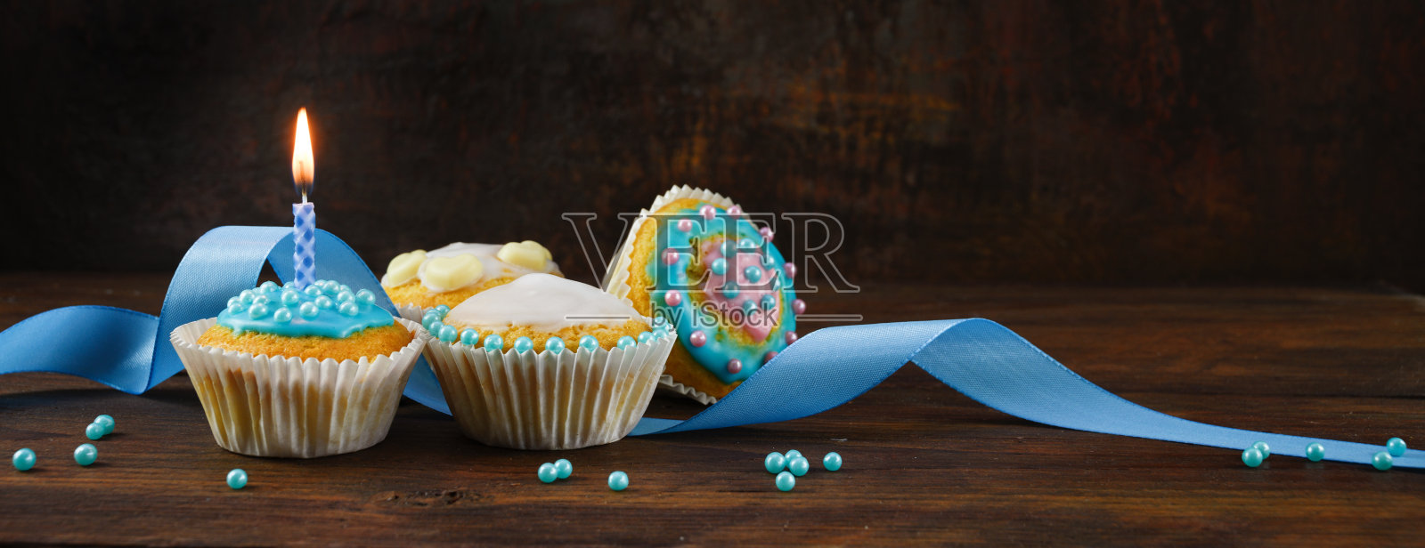 装饰松饼或纸杯蛋糕与蓝色丝带和燃烧的蜡烛在一个质朴的木板上，以暗棕色的背景，全景格式与复制空间照片摄影图片