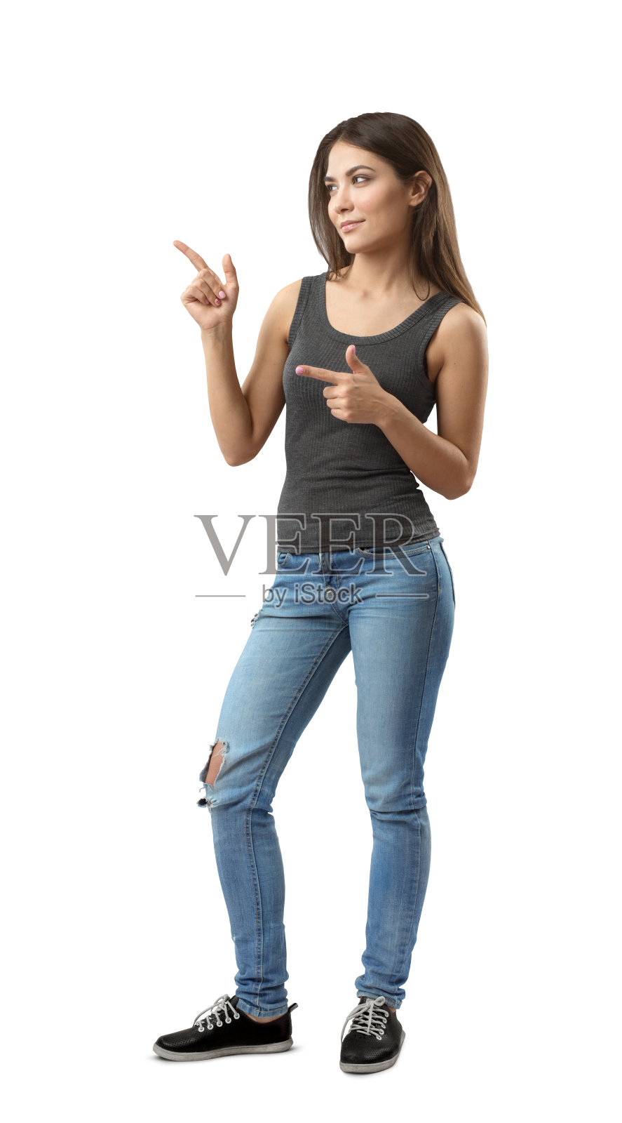 穿着无袖上衣和牛仔裤的年轻女子半转身站着，双手弯曲，食指指向旁边，孤立在白色背景上。照片摄影图片