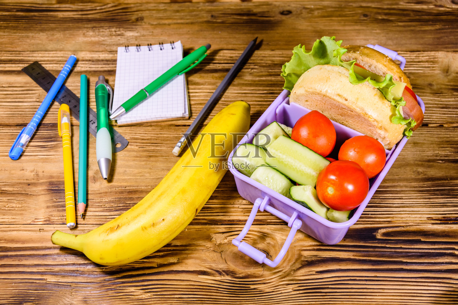 香蕉，不同的文具和装有汉堡、黄瓜和西红柿的午餐盒放在木桌上照片摄影图片