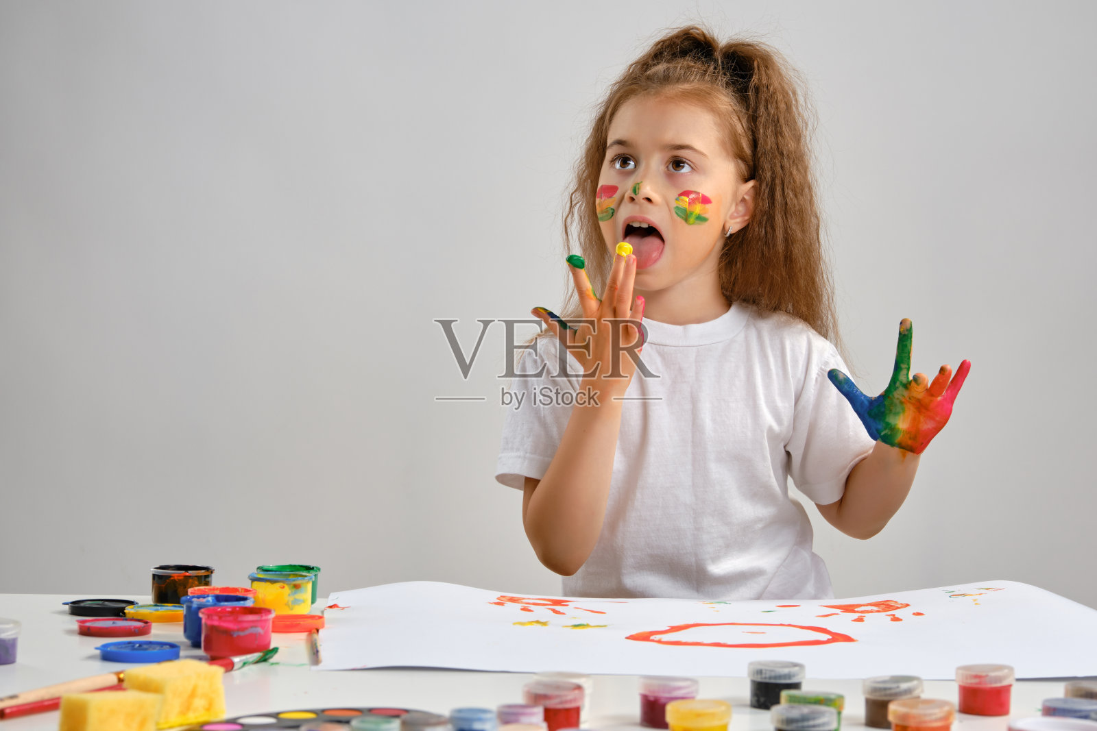 一个穿着白色t恤的小女孩坐在桌子旁边，桌子上画着什么，舔着手上的颜料。孤立在白色的。媒介特写照片摄影图片