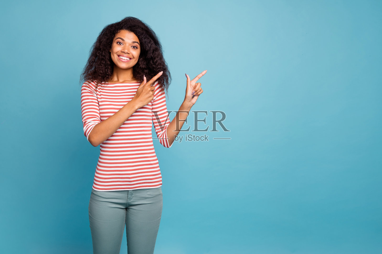 开朗可爱可爱迷人甜美漂亮的年轻人指着空穿灰色裤子的裤子孤立在蓝色鲜艳的背景照片摄影图片