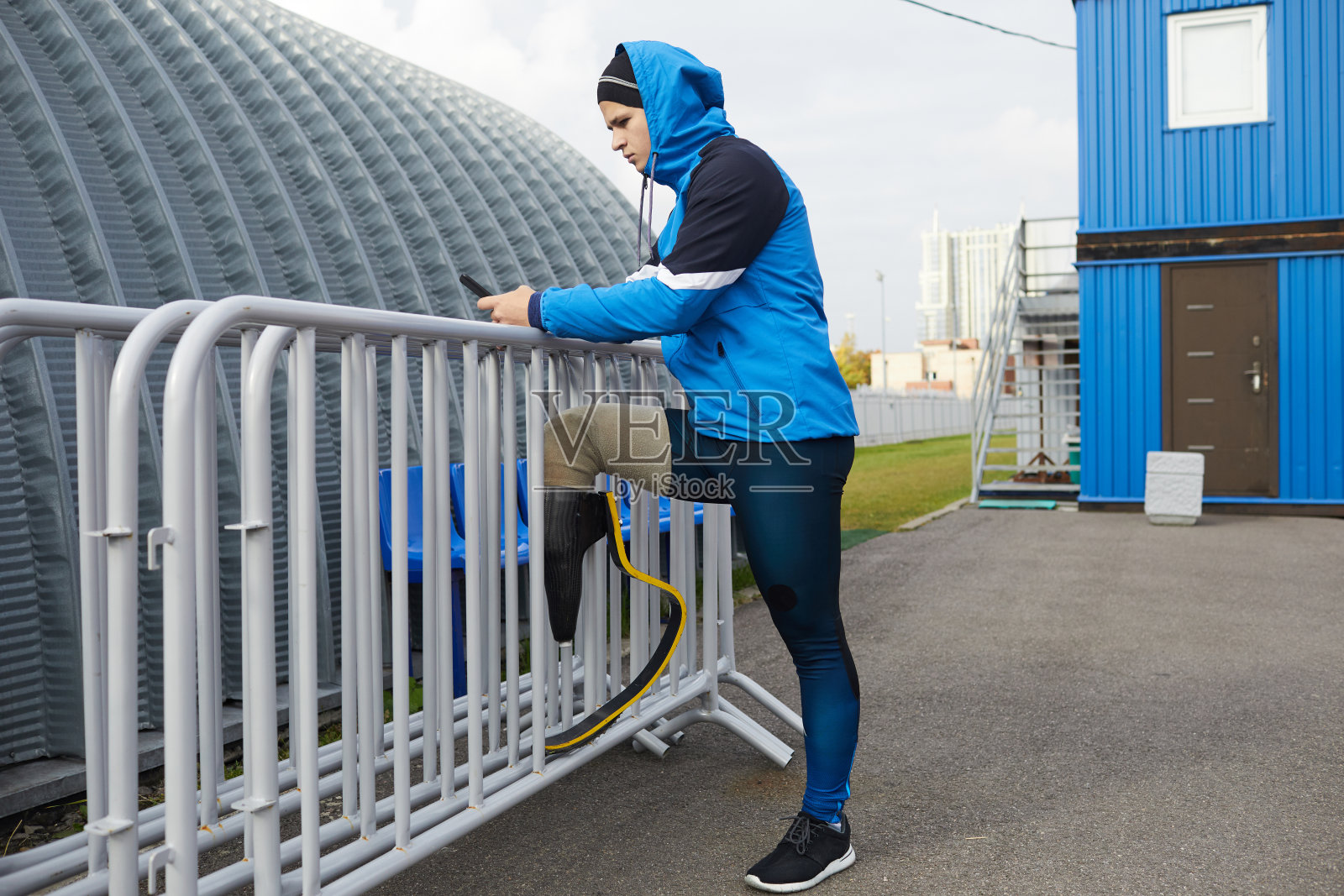 严肃的年轻截肢运动员穿着夹克把假肢装在栏杆上，在体育场浪费时间使用电子设备照片摄影图片