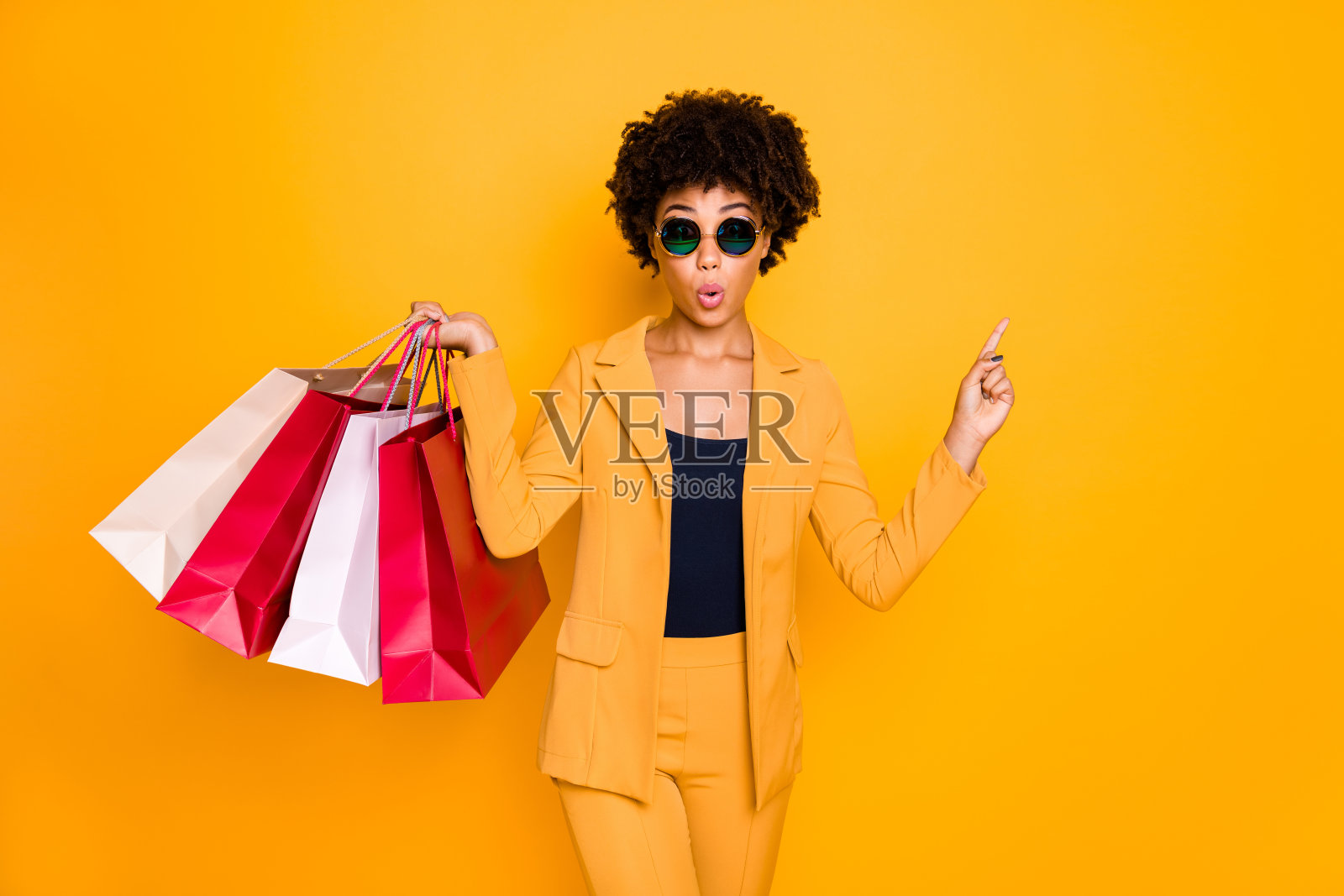 看起来令人难以置信的销售。惊讶的黑发卷发女孩点食指copyspace购物跟随秋天的折扣拿包穿夹克裤子孤立的黄色背景照片摄影图片