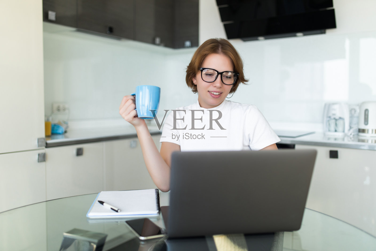 一个年轻的女人拿着咖啡看着她面前的笔记本电脑在厨房里照片摄影图片