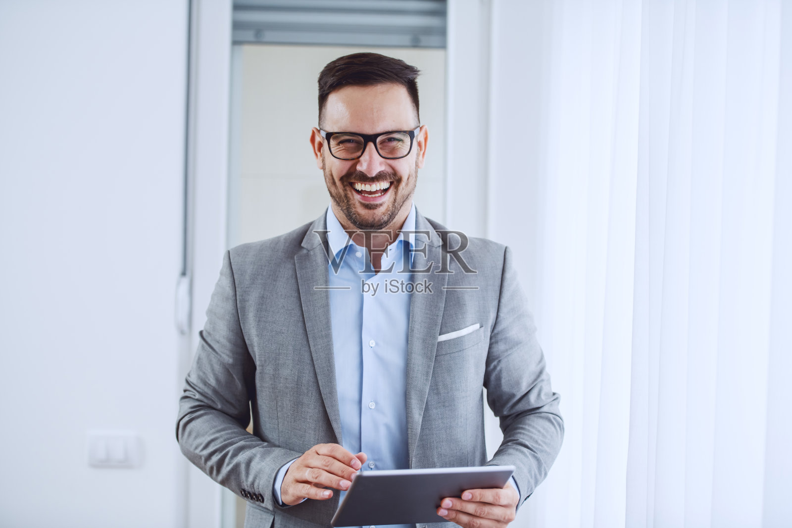 漂亮开朗的白人优雅的商人穿着西装，戴着眼镜，拿着平板电脑，站在窗户旁边看着相机。照片摄影图片