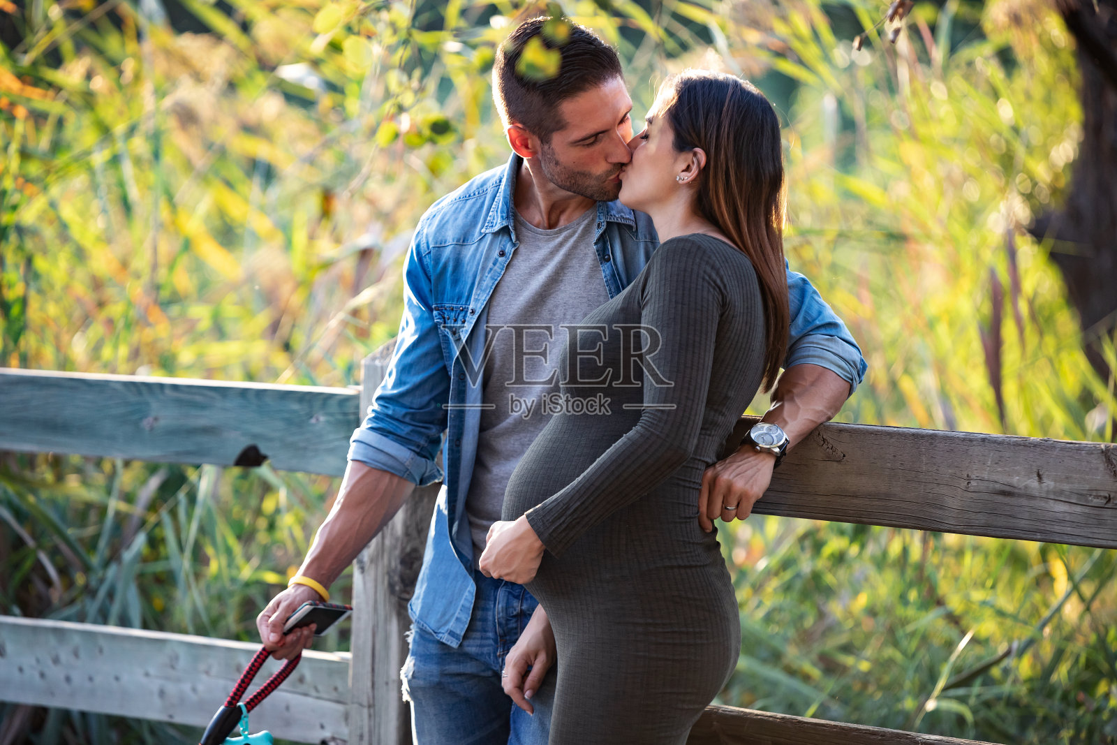 阳光明媚的公园里，时尚孕妇夫妇在秋叶下亲吻。照片摄影图片