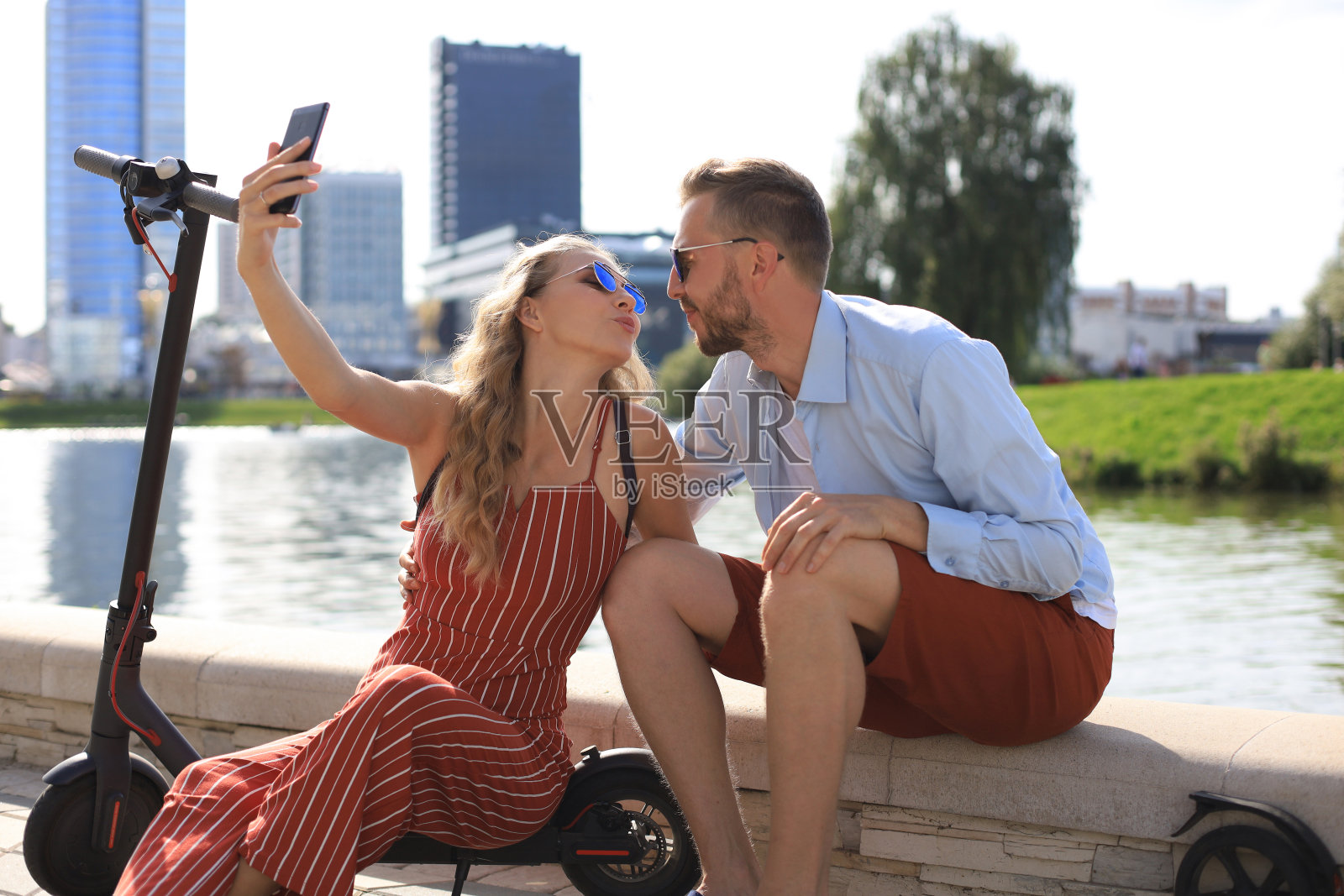 可爱的夫妇驾驶电动滑板车，从驾驶中休息，坐在河岸上，自拍照片摄影图片