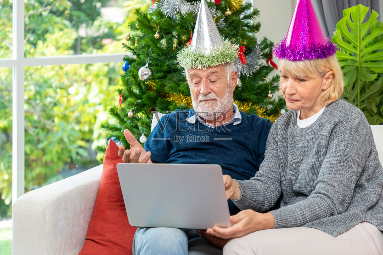 一对老年夫妇坐在装饰有圣诞树的沙发上，用笔记本电脑视频打电话问候他们的家人过圣诞节照片摄影图片