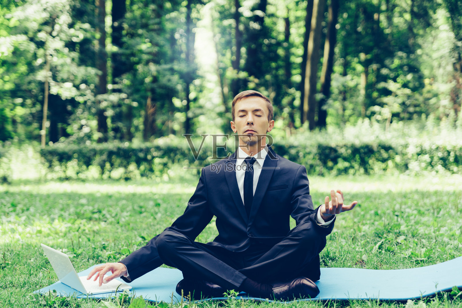 穿西装打领带的男人。一个商人在公园里以莲花的姿势休息，他不能以任何方式放松，用一只手在电脑上打字。照片摄影图片