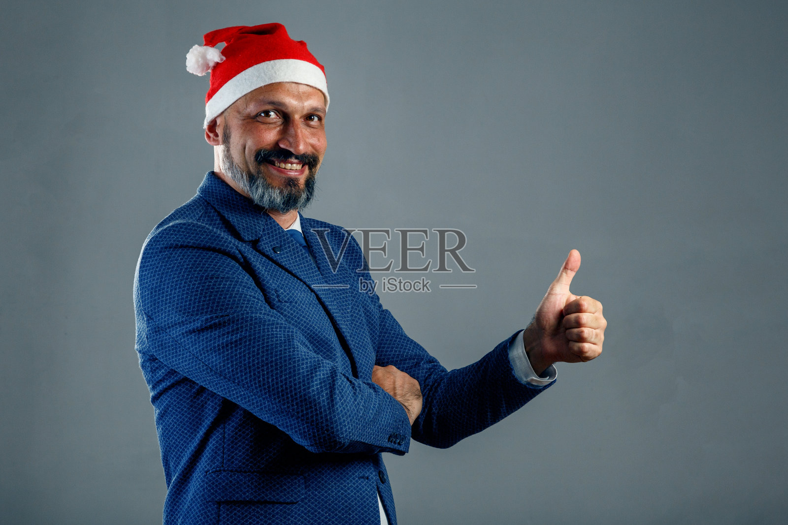 一名身穿蓝色夹克、头戴新年帽的大胡子男子站在灰色背景上，做出表示赞同的手势照片摄影图片