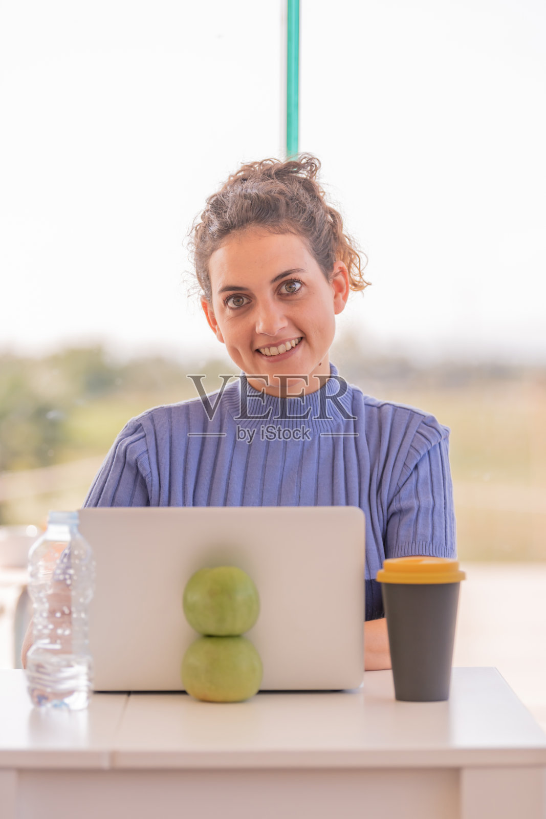 一个友好的女孩坐在一张桌子前看着相机，桌子前有一台白色的笔记本电脑，上面有两个绿苹果，背景没有对焦。照片摄影图片