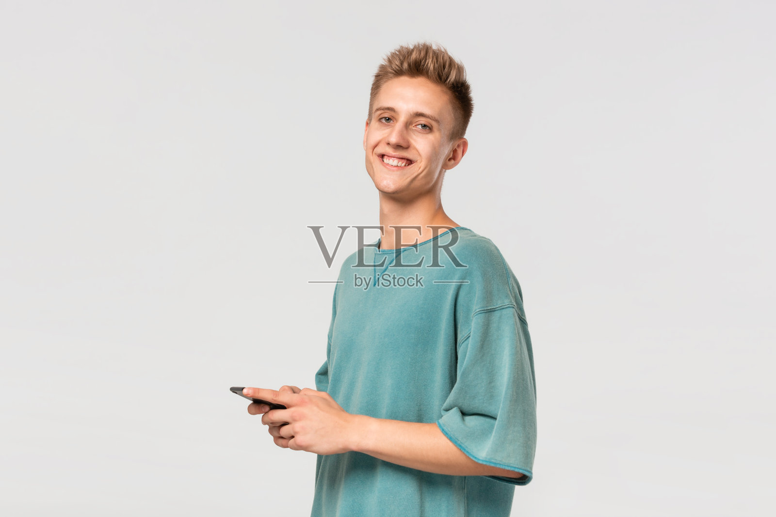 穿着绿色t恤的金发少年微笑着，拿着智能手机，看着淡灰色背景下孤立的摄像头。照片摄影图片