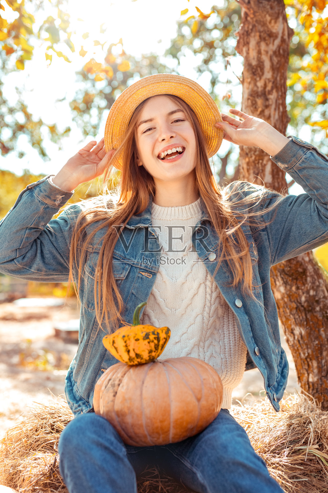 万圣节Preparaton概念。年轻女子穿着牛仔装坐在花园里，拿着南瓜收获帽摆姿势笑得开心照片摄影图片