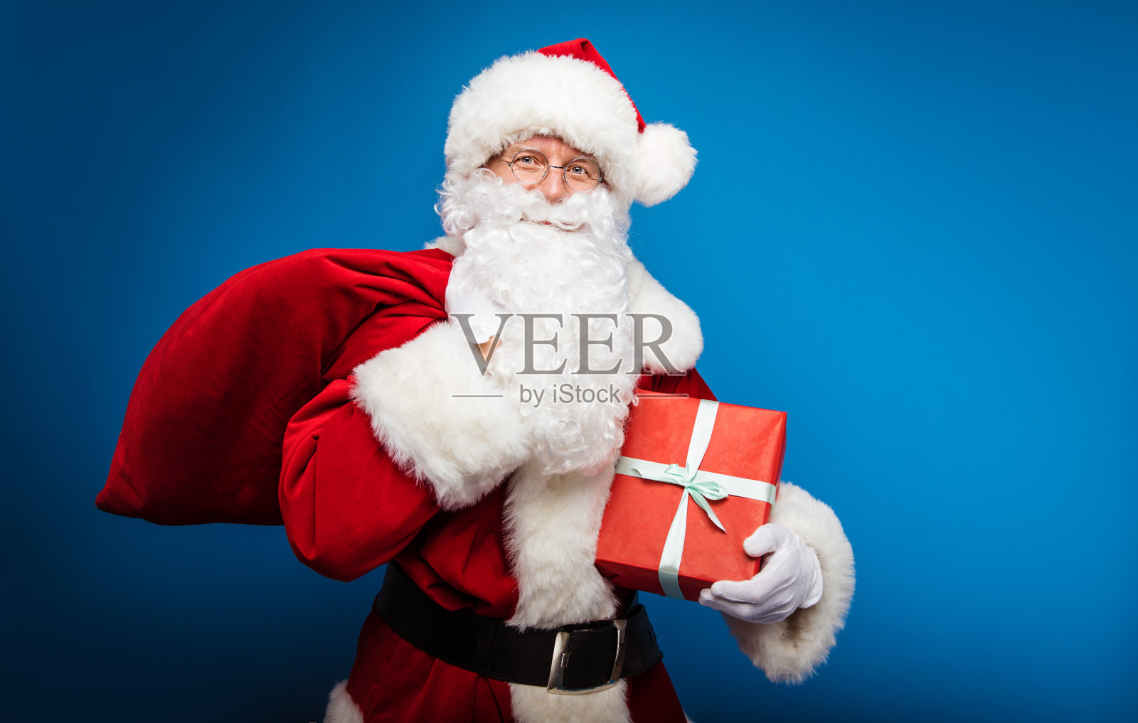 孩子们的英雄。真正的圣诞老人正摆姿势，右肩上扛着一袋礼物，右臂上挎着一份用红色包装纸包装的礼物，看着镜头。照片摄影图片