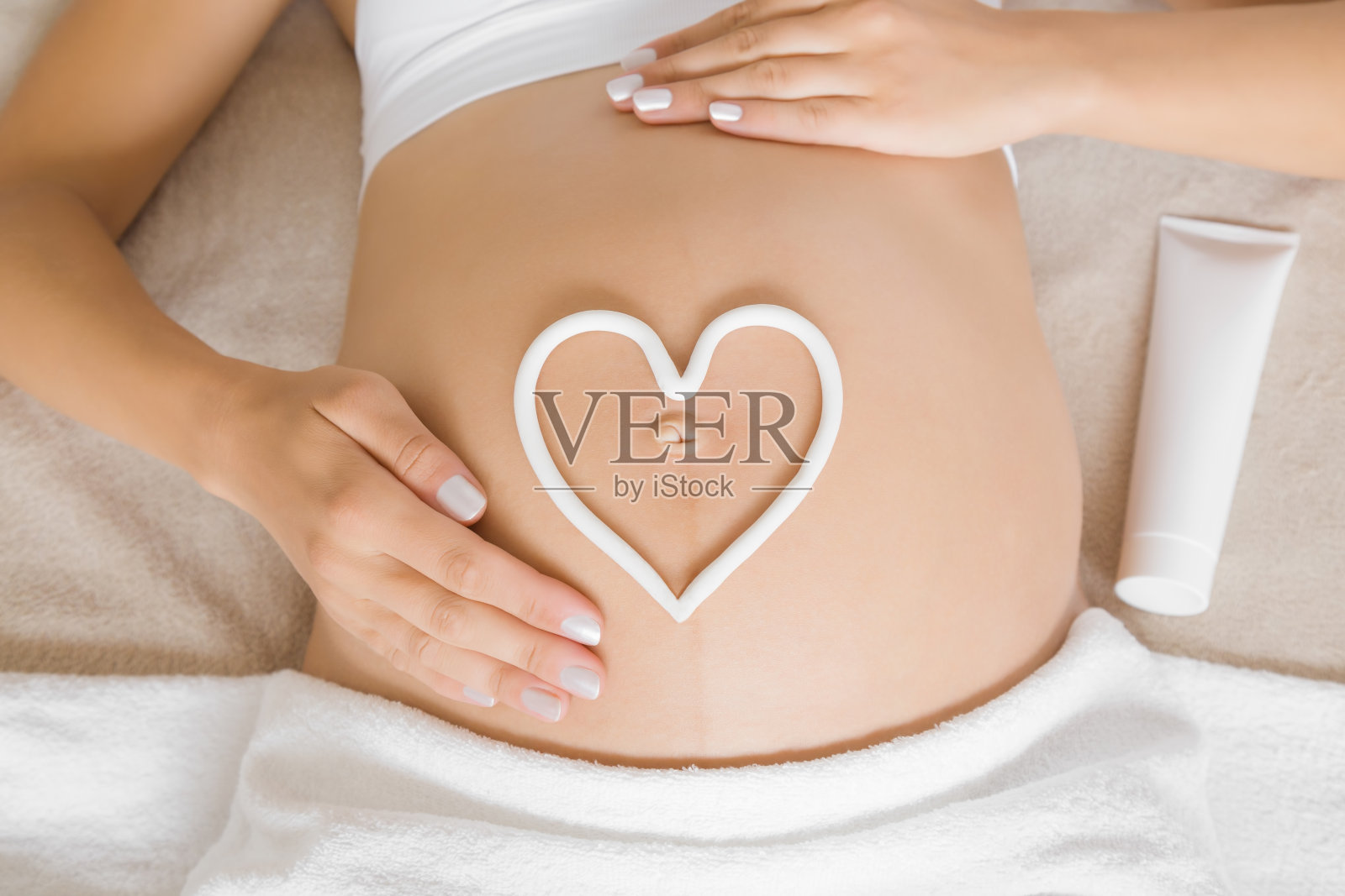 年轻女子躺在床上。温柔的手触摸。白色的心形是在裸露的大肚子上用奶油做成的。呵护怀孕期间肌肤的完美光滑。关闭了。前视图。照片摄影图片