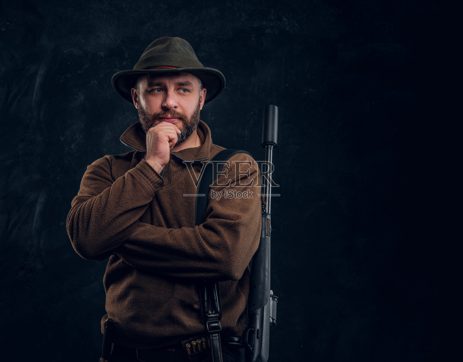 一个蓄着胡须的猎人的肖像，手握步枪，下巴上带着沉思的神情。暗墙背景下的工作室照片照片摄影图片