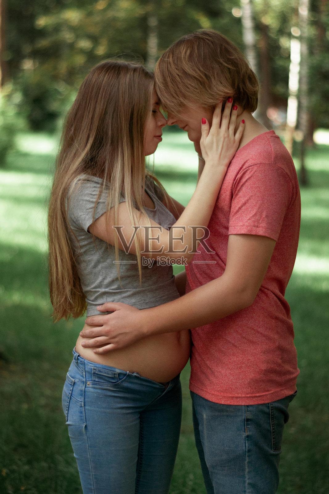 竖版照片：幸福的已婚夫妇，丈夫拥抱怀孕的妻子，在公园里散步，孕中期，手放在妻子的肚子上照片摄影图片