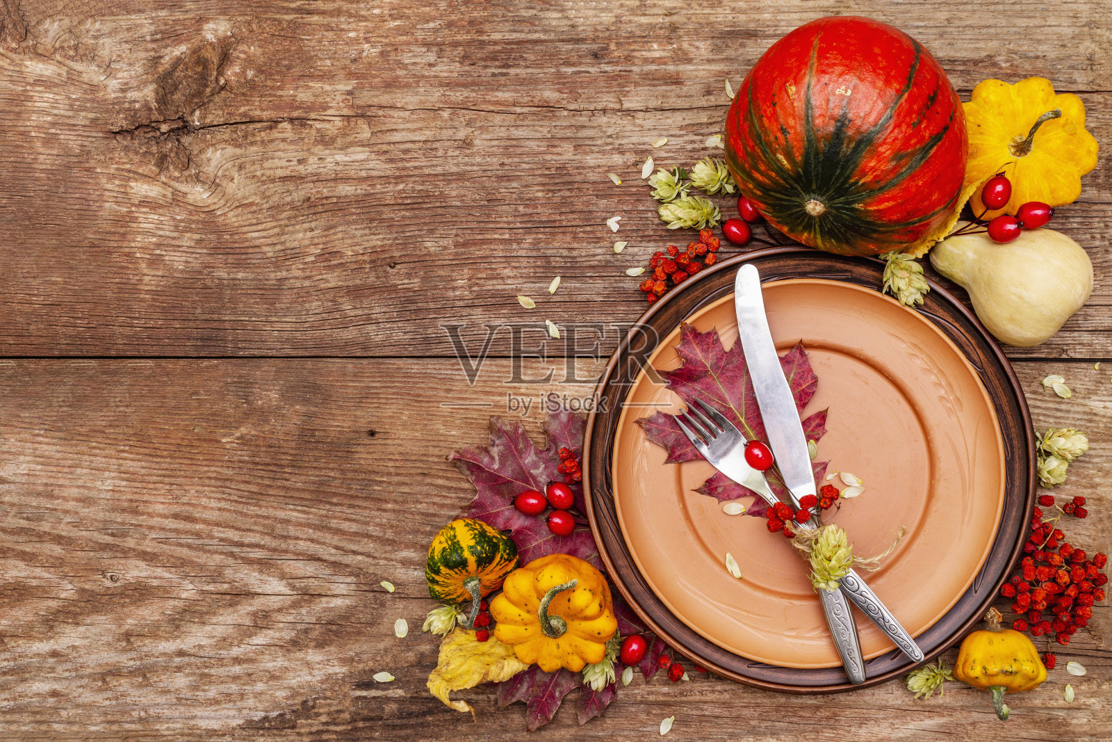秋天和感恩节晚餐的地方设置。收获的蔬菜和浆果，五颜六色的叶子，啤酒花，陶瓷盘子，叉子和刀。秋季餐具木制背景照片摄影图片