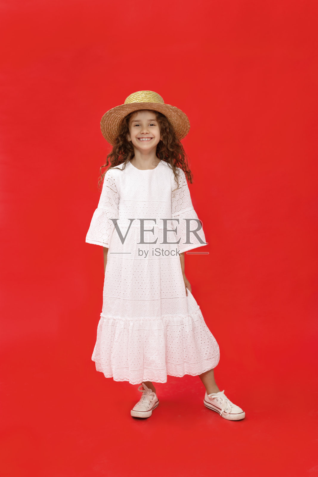 快乐的小卷女孩，穿着白色的衣服和帽子，看着相机，在红色的背景。垂直的观点。照片摄影图片