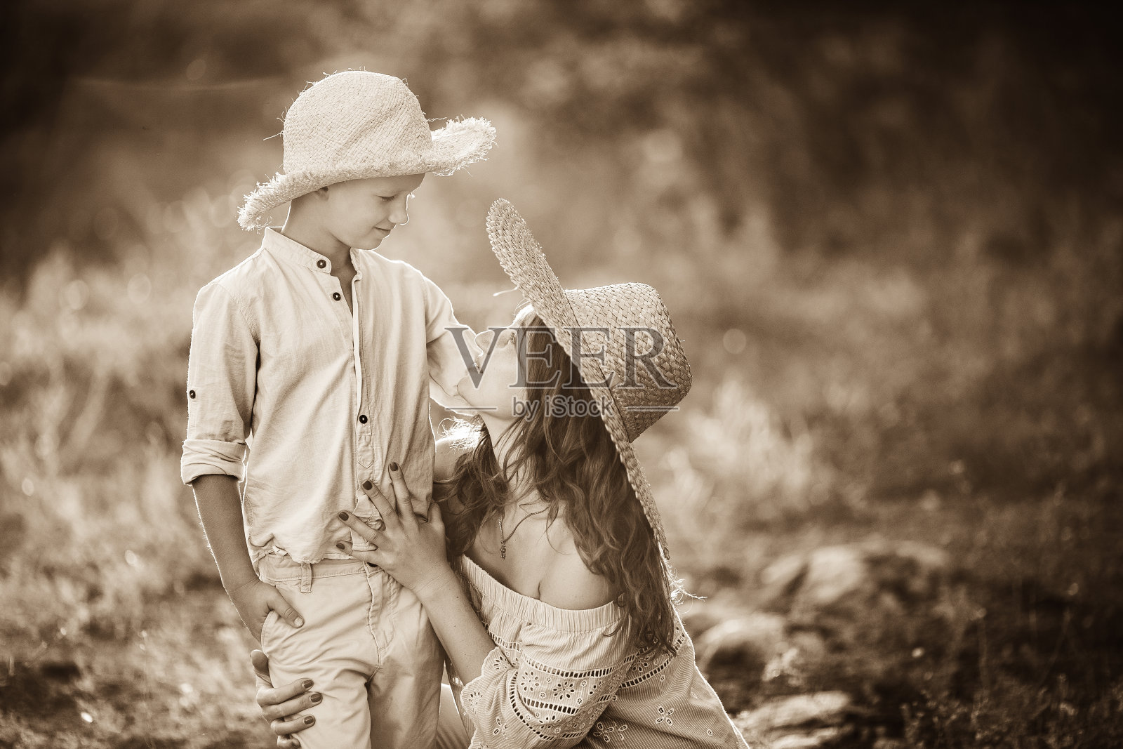 落日背景下的红发女孩带着一个孩子。有个金发宝宝的可爱妈妈。妈妈和戴草帽的儿子在一起照片摄影图片