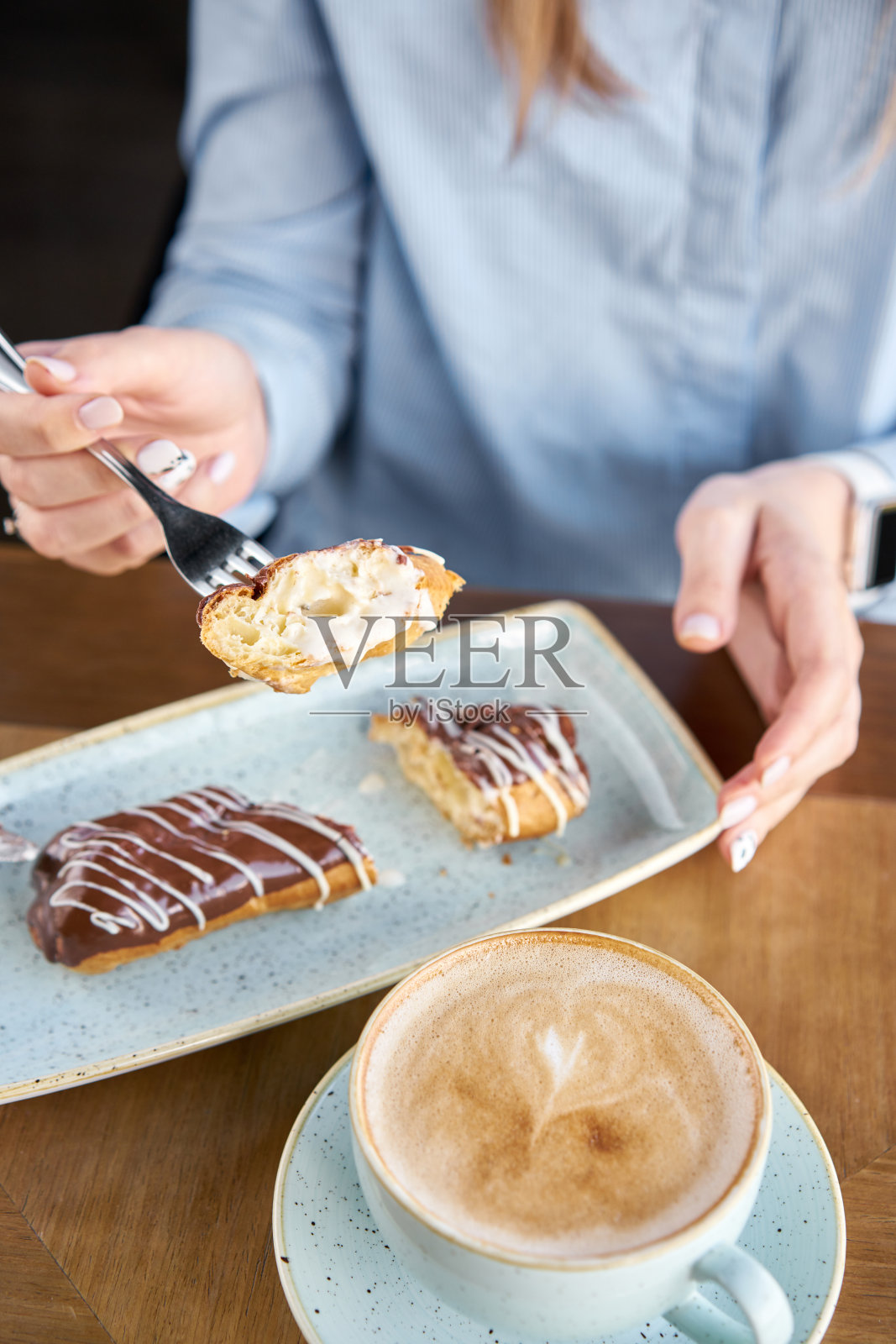 ． 传统的法式巧克力泡芙。一个年轻的女人在餐厅喝咖啡卡布奇诺吃甜点。用香草奶油和巧克力糖霜装饰的蛋糕。照片摄影图片