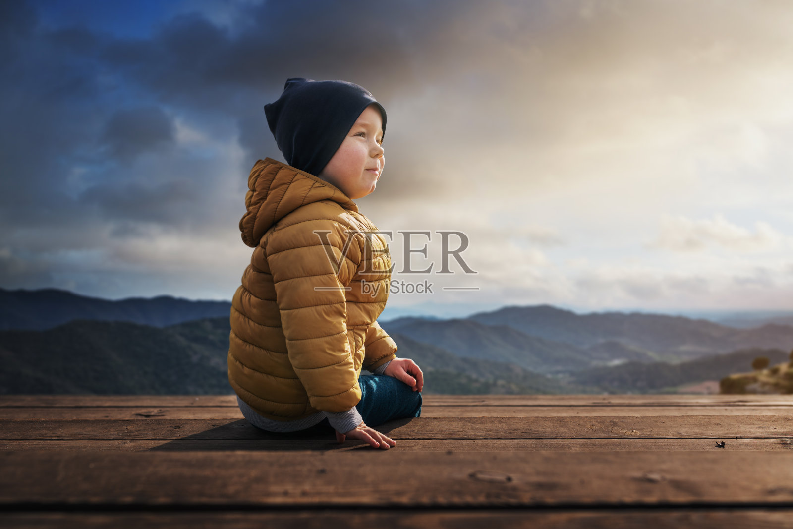 日落时，小男孩坐在木地板上。美丽的风景作为背景。照片摄影图片