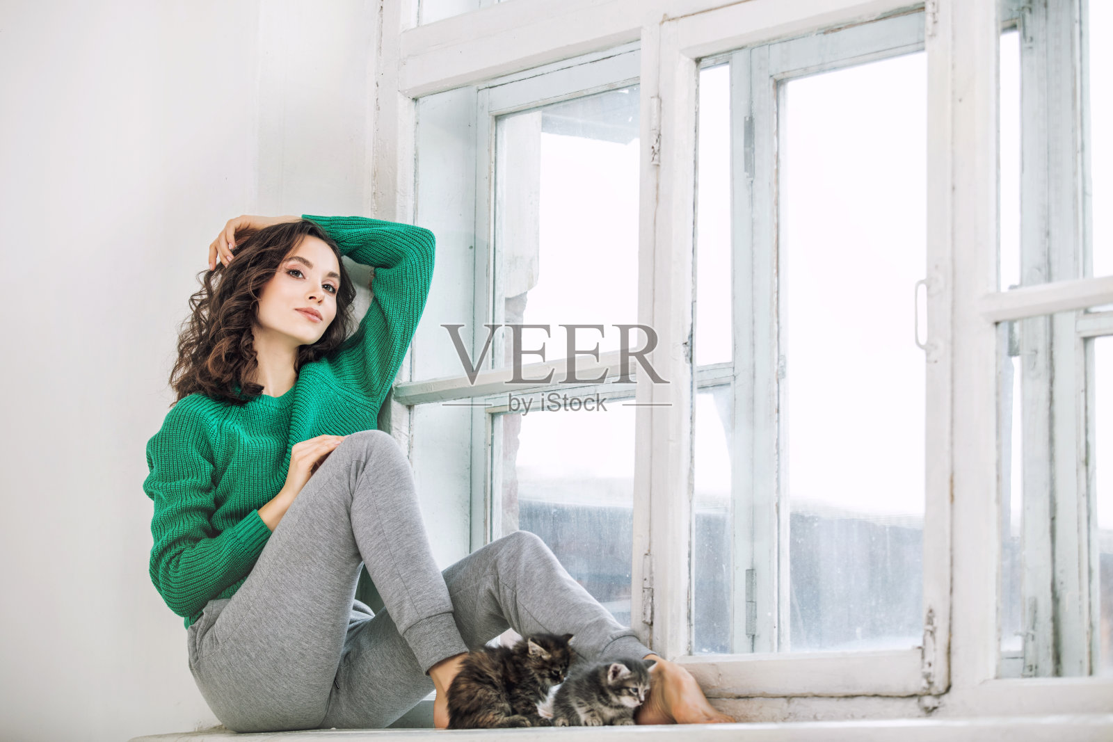 穿着毛衣的年轻漂亮女孩坐在白色窗台上和小猫们在一起照片摄影图片