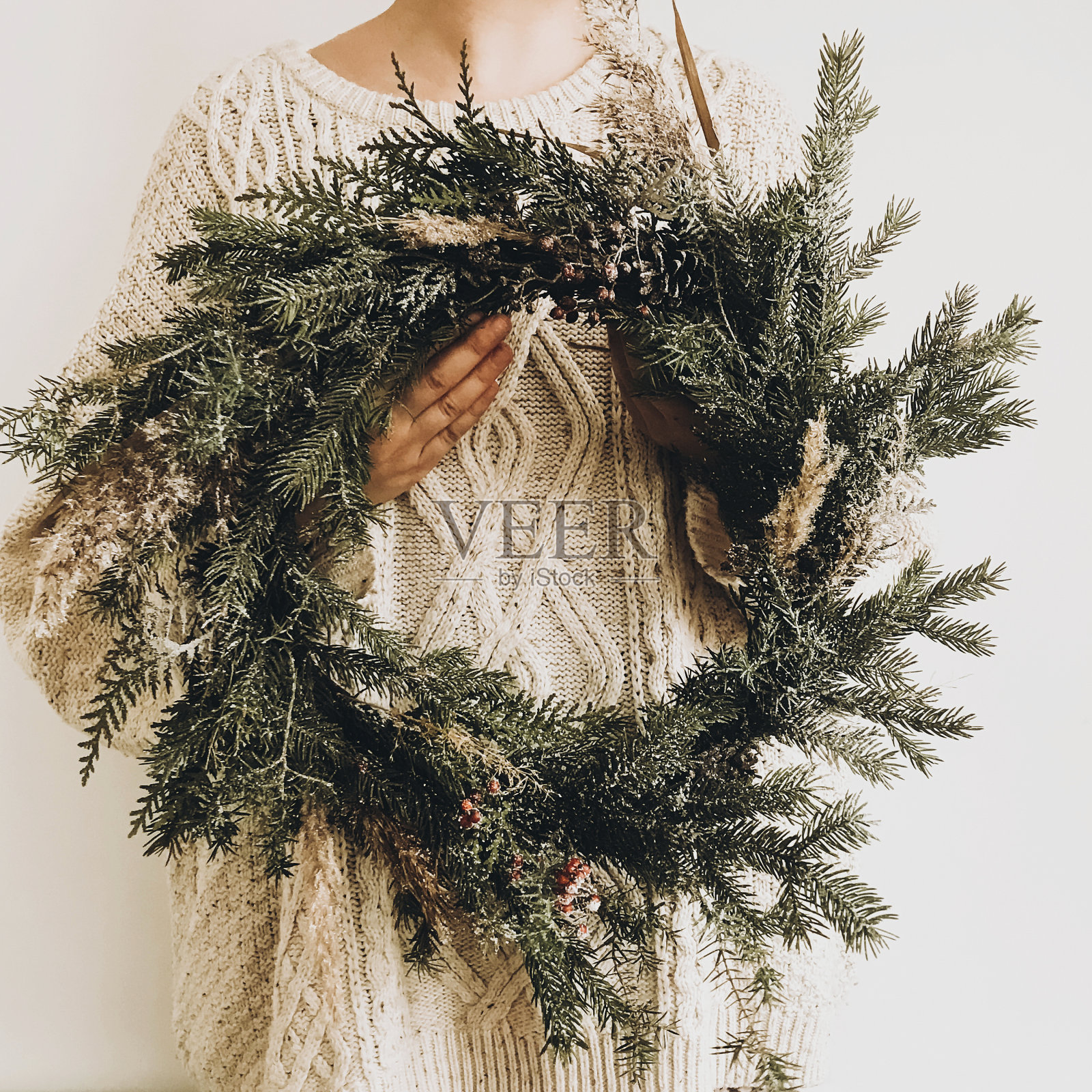 女孩手里拿着时髦的乡村花环。嬉皮士女孩在针织毛衣抱着农村现代花环在白色的墙壁背景。节日装饰。手机照片照片摄影图片