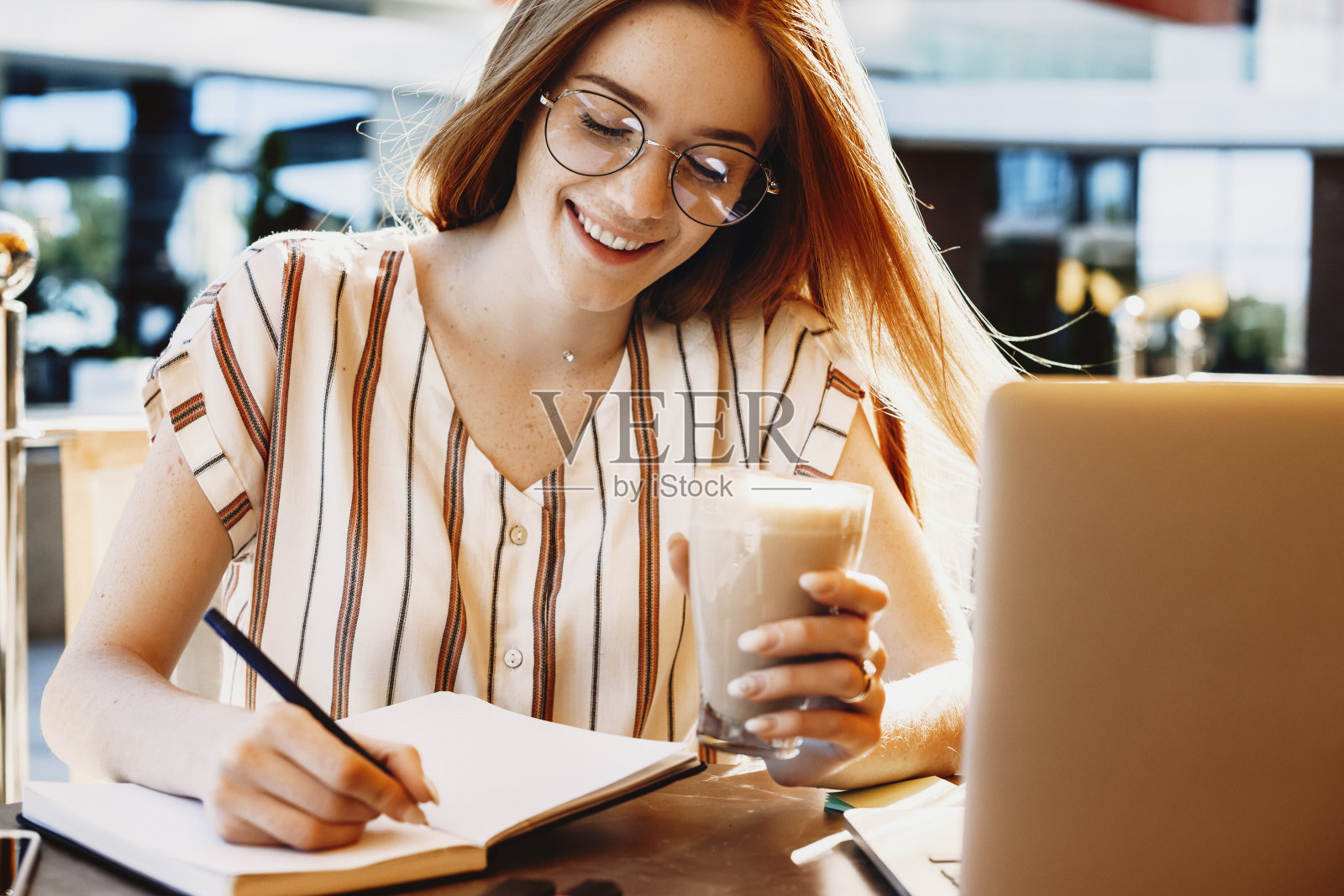 一个可爱的年轻女子的特写笔记，而自由职业者在咖啡店外面。可爱的红发雀斑女正在外面做视频。照片摄影图片