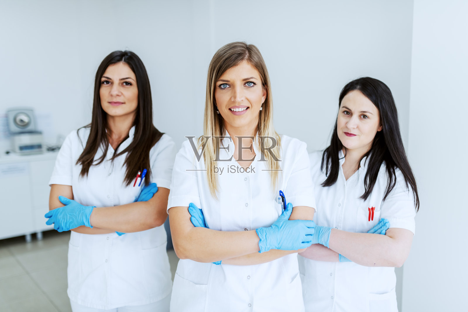 三个漂亮的白种人实验室助理穿着白色制服，戴着橡胶手套，双手交叉站在实验室里。照片摄影图片