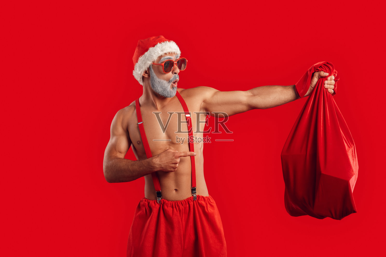 圣诞节自由泳。年轻的大胡子圣诞老人赤裸上身，戴着帽子和太阳镜，孤立地站在红色的礼物袋指着一边惊讶照片摄影图片