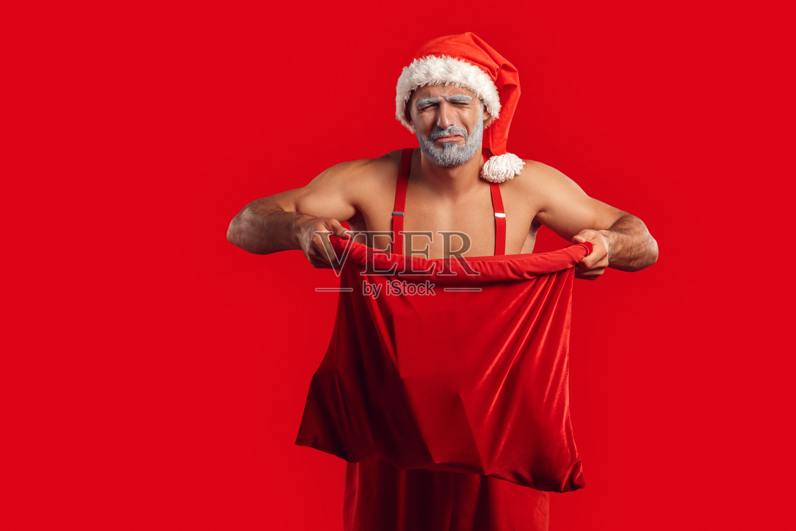 圣诞节自由泳。年轻的圣诞老人裸露着肌肉的上半身，在帽子上孤立地站在红色的打开的礼物袋上，苦笑着失望照片摄影图片