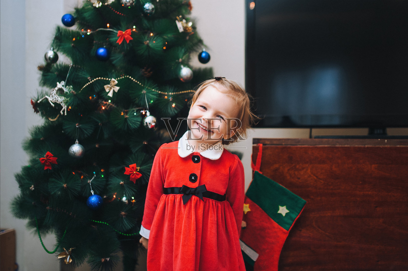 漂亮可爱的小女孩装扮成圣诞老人。在装饰好的圣诞树的背景上，她讲着一首新年诗，愉快地微笑着。照片摄影图片