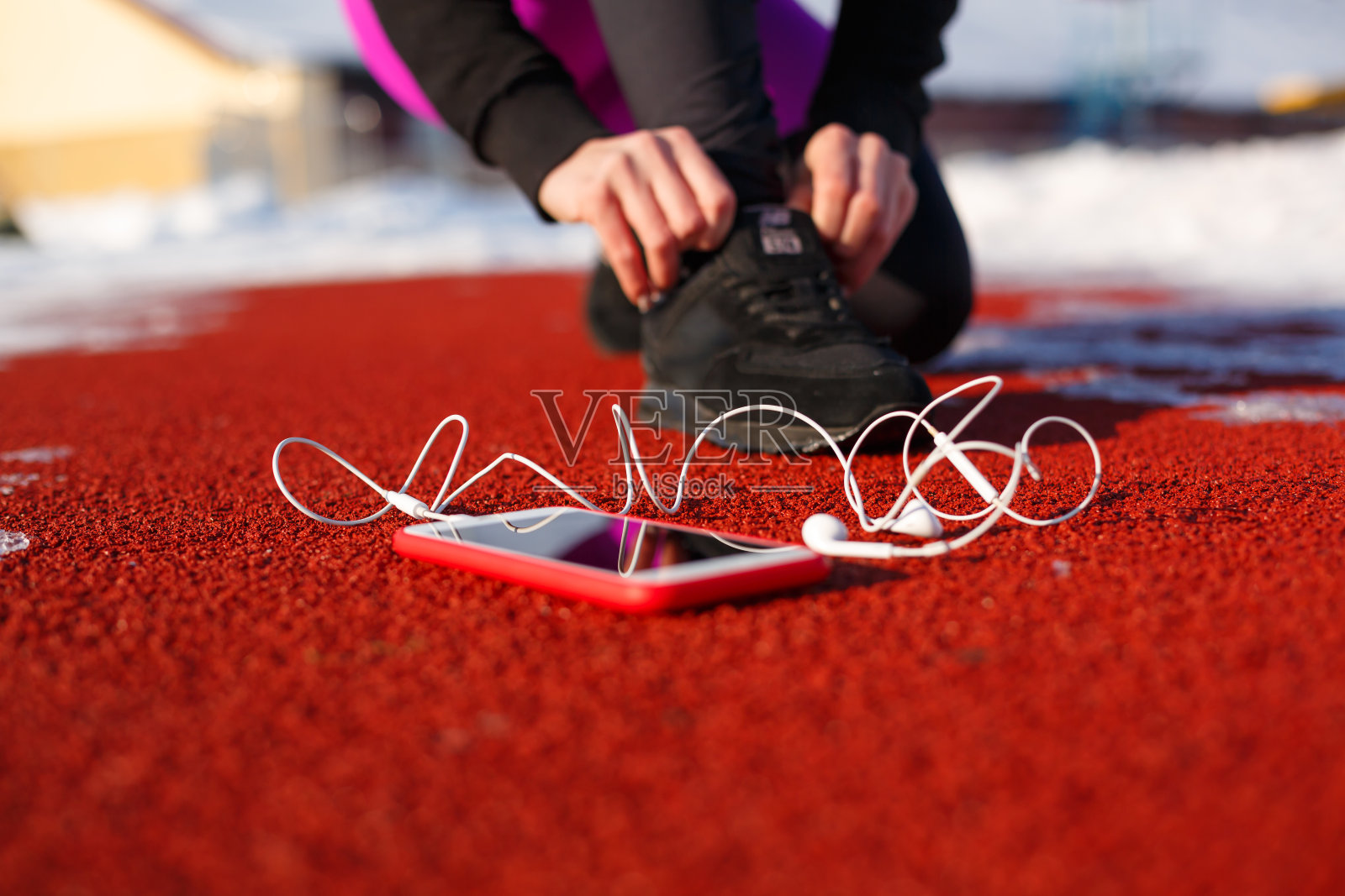 穿着黑色运动鞋的女运动员，蹲在红色的跑道上跑步。附近有一部带有线耳机的电话。寒冷的天气照片摄影图片