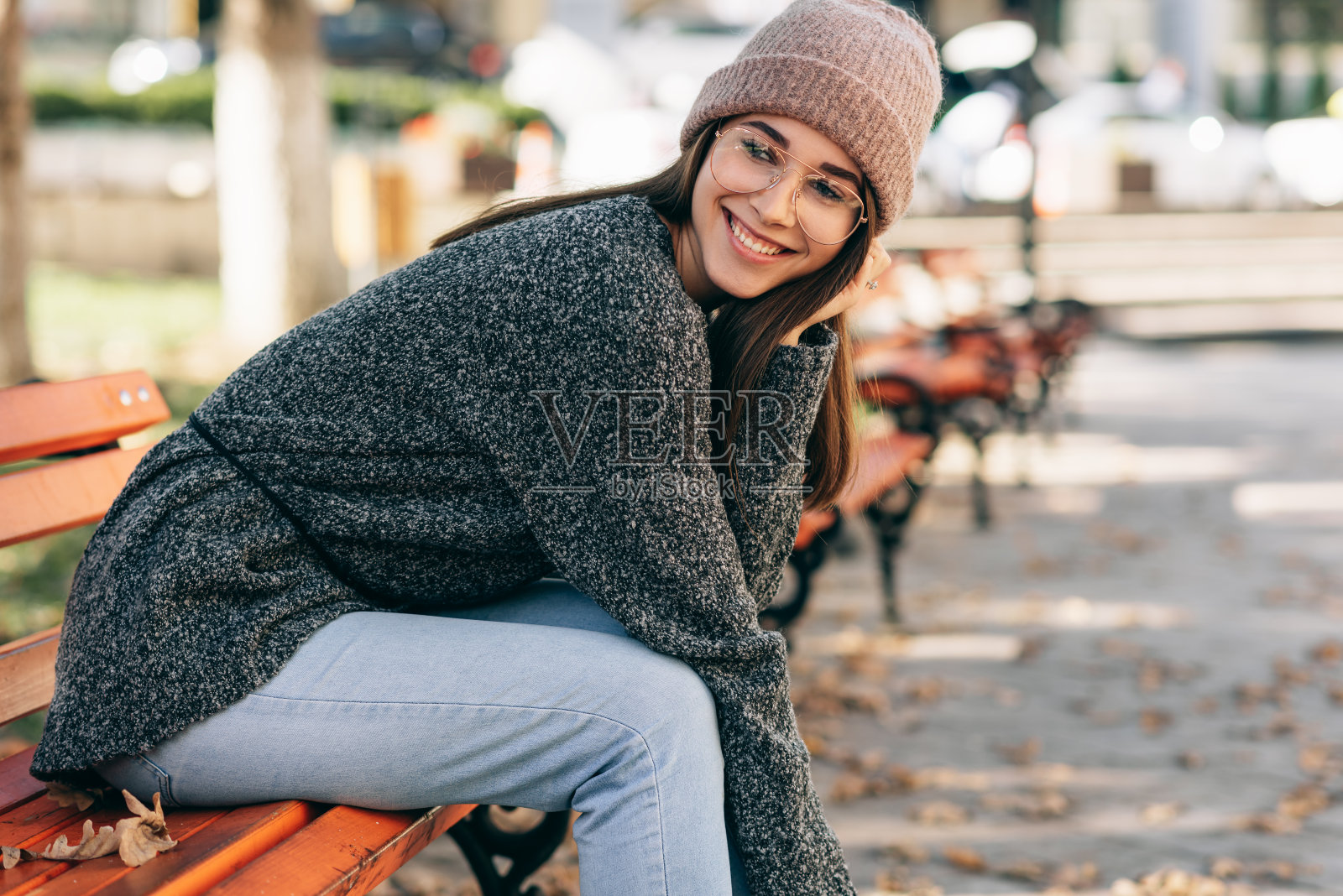 这张照片里的女学生面带微笑，看着镜头，穿着针织毛衣，戴着眼镜和帽子，坐在城市街道的长凳上。漂亮的年轻女子在外面休息照片摄影图片