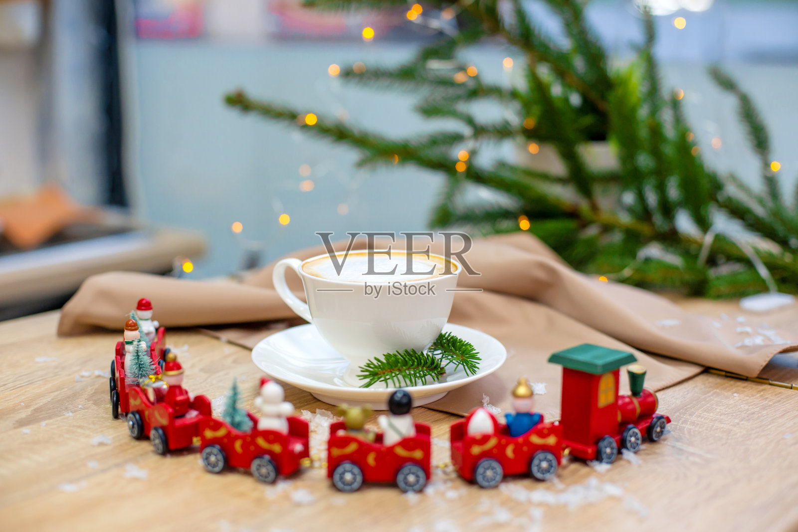 美味新鲜的节日早晨卡布奇诺咖啡装在陶瓷白色的杯子里，放在装饰圣诞的火车、红色的装饰物、萤火虫和云杉树枝的木桌上。照片摄影图片